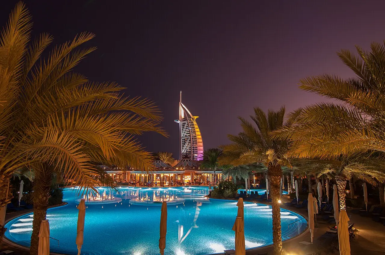 Burj Al Arab's sail-shaped tower glowing at night beyond palm trees and a lit pool